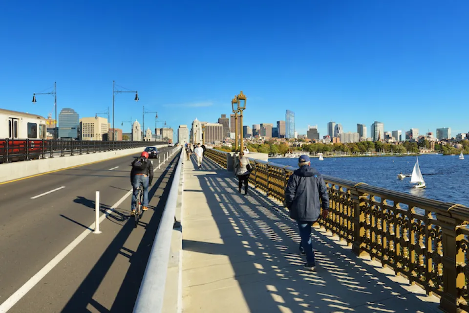 View of Boston skyline from Longfellow Bridge with cyclist riding in bike lane and pedestrian on sidewalk.